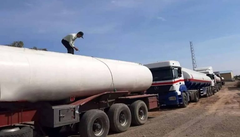 An Iranian man stands on top of a large white fuel tanker truck in a queue with tankers transporting fuel