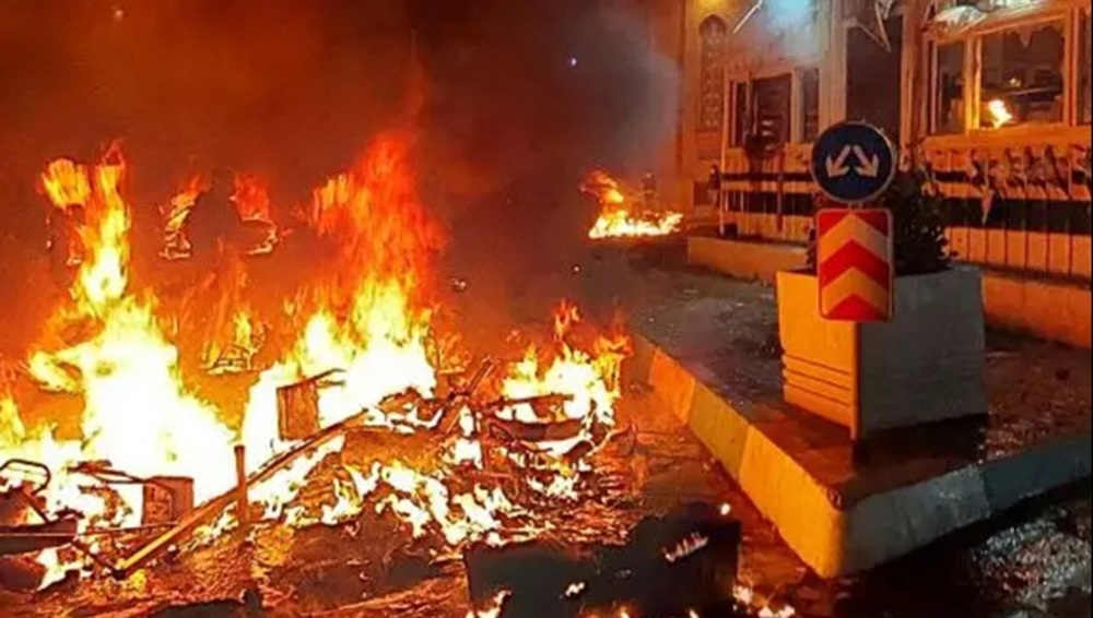 Iranian protesters block a city street at night by burning piles of debris and trash bins.