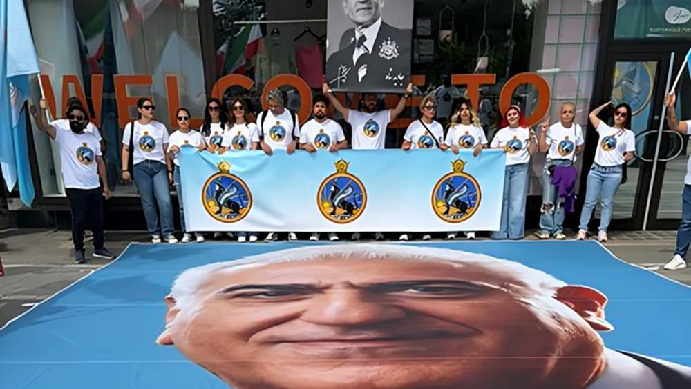 Iranian monarchist supporters in Regensburg, Germany, May 10, 2026, pose with oversized portraits of Reza Pahlavi (foreground) and the ousted Shah (held aloft), displaying SAVAK banners and emblem T-shirts