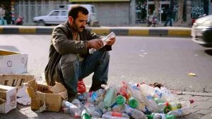 Man crouching on a city street in Iran, sorting through a pile of discarded plastic bottles to scrape together a basic livelihood