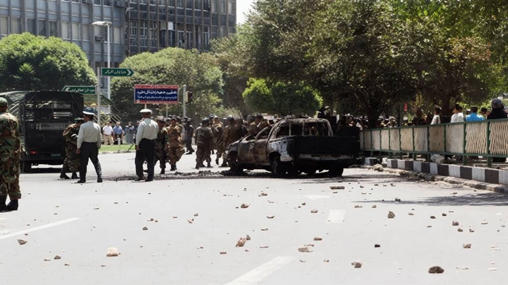 Security forces stand near a burnt-out vehicle amidst a street littered with debris following clashes in Tehran during the July 1999 student uprising