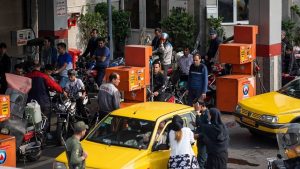 Crowds of people, motorcycles, and taxis line up at a busy gas station in Iran as drivers wait for fuel