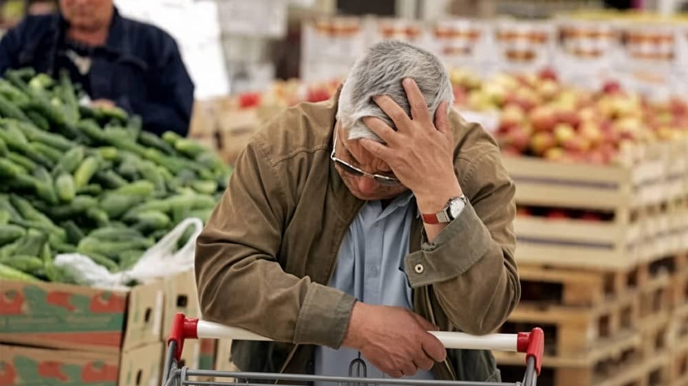 An elderly Iranian man rests his head in his hand while leaning on a shopping cart, his posture and expression conveying exhaustion and quiet distress