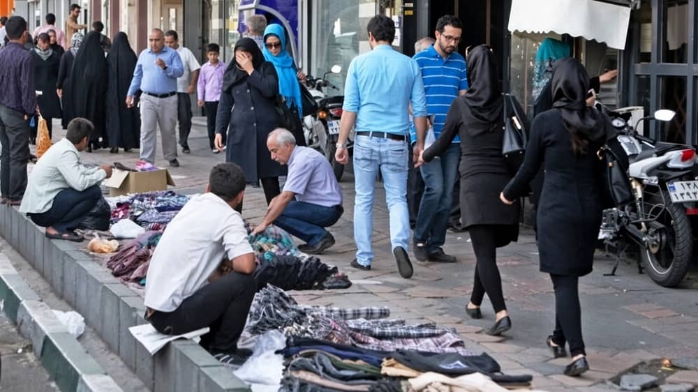 Street vendors display clothes on the sidewalk as shoppers stroll past storefronts in a busy Iranian city market