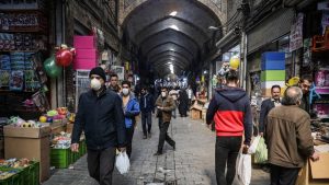 A quiet current of daily life moves through the vaulted corridors of an old covered bazaar, where shoppers weave past toy stalls, hanging balloons, and stacked crates under rows of hanging lamps.
