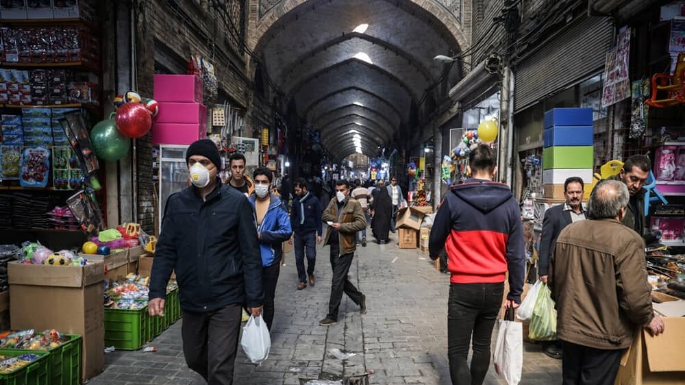 A quiet current of daily life moves through the vaulted corridors of an old covered bazaar, where shoppers weave past toy stalls, hanging balloons, and stacked crates under rows of hanging lamps.